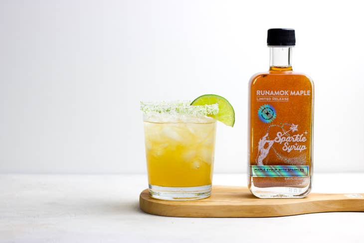 Sparkle maple syrup bottle beside a glass with yellow drink and lime wedge on board.