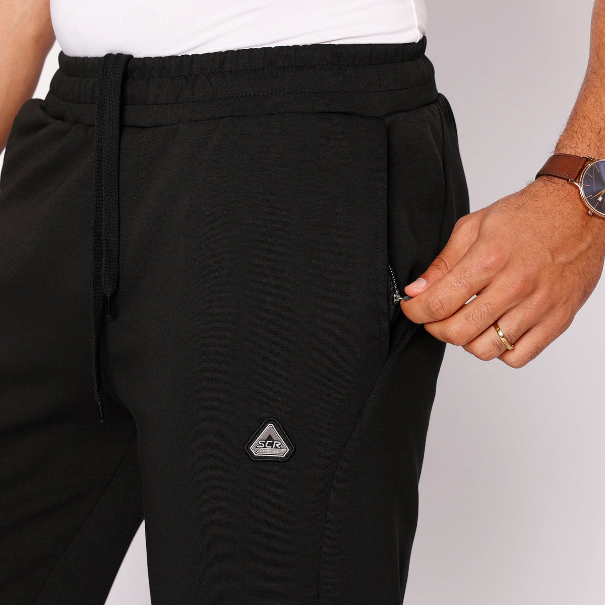 Black sweatpants close-up: elastic waistband, drawstring, SCR logo patch on leg.