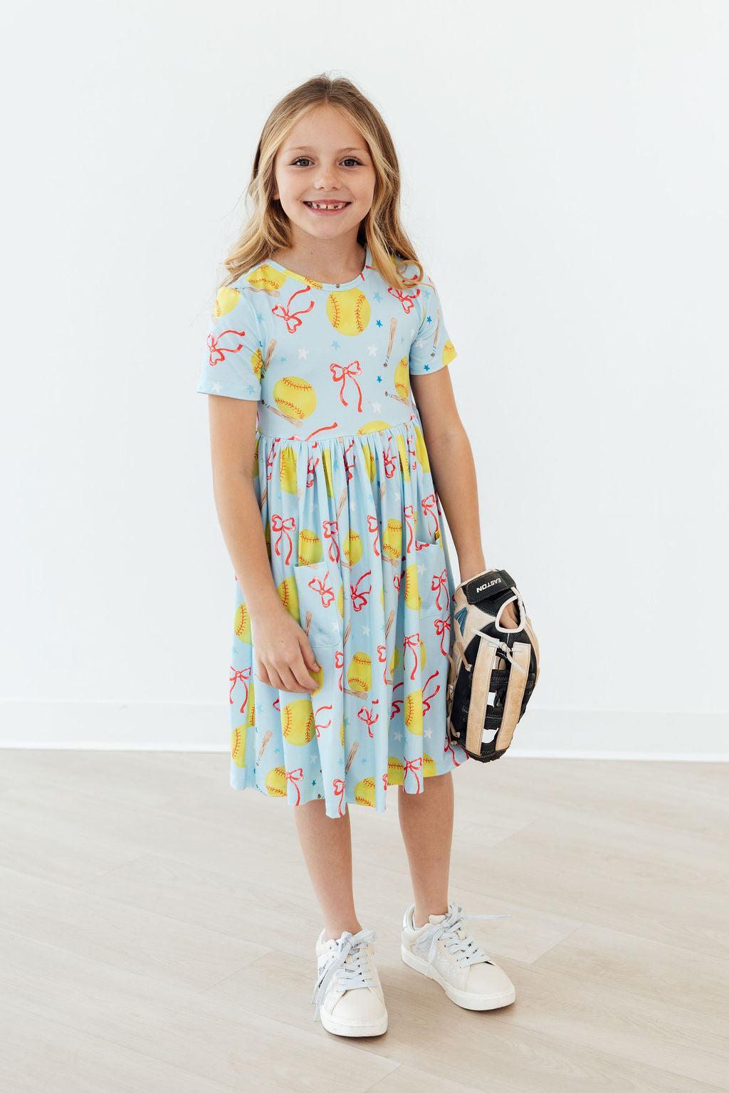 Light blue short-sleeve twirl dress with colorful softball and bow print, holding a glove