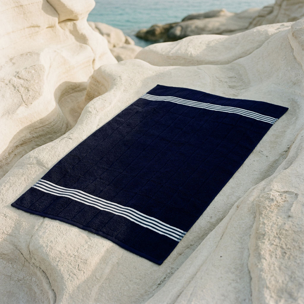 Navy Australian cotton bath towel with white striped border, laid on rocks