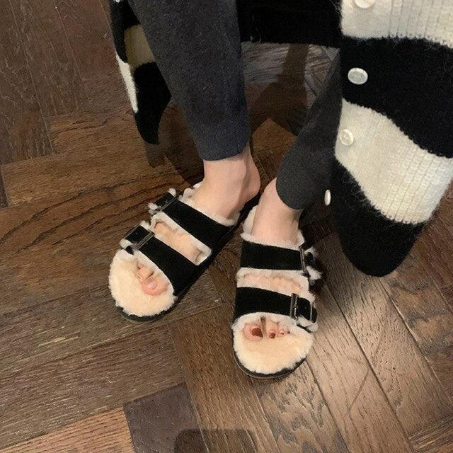Black double-buckle faux-fur slippers on wooden floor, cozy indoor outdoor design