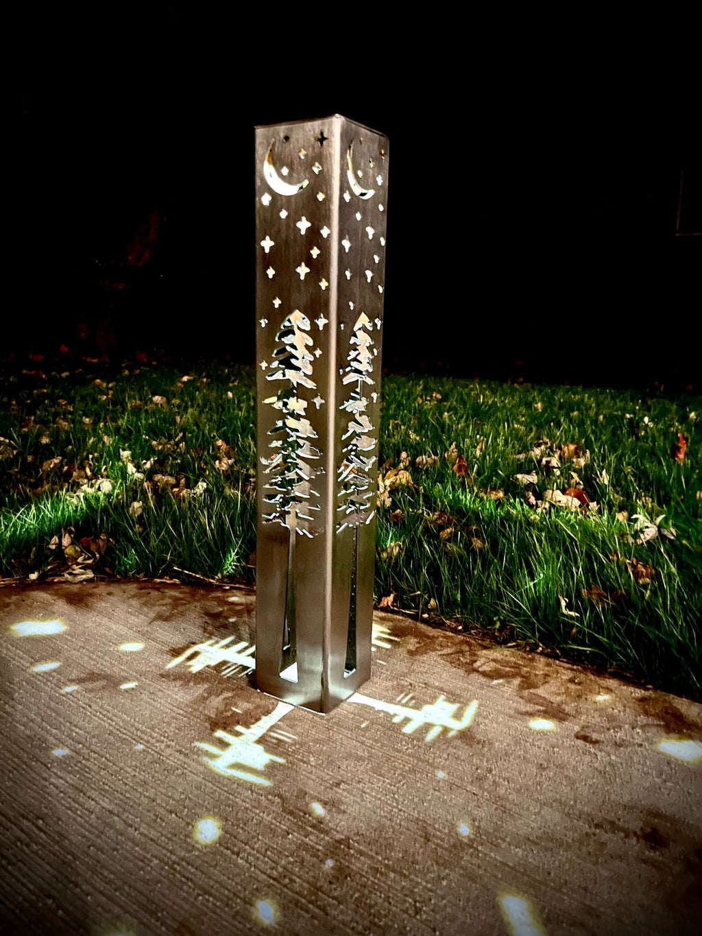 Solar light pillar standing in grass at night, casting star and pine tree light patterns.