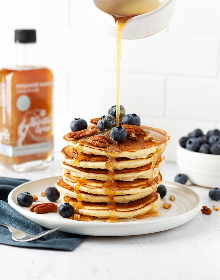 Sparkle maple syrup bottle pouring golden syrup over pancakes with blueberries.