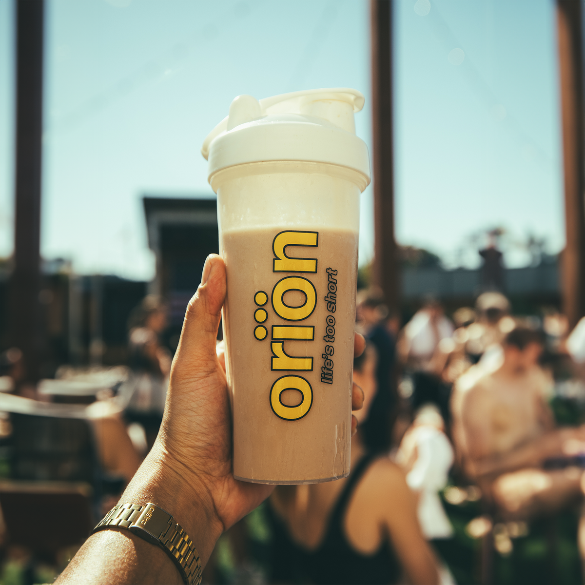 Hand holding white protein shaker with gold Orion logo and light brown drink