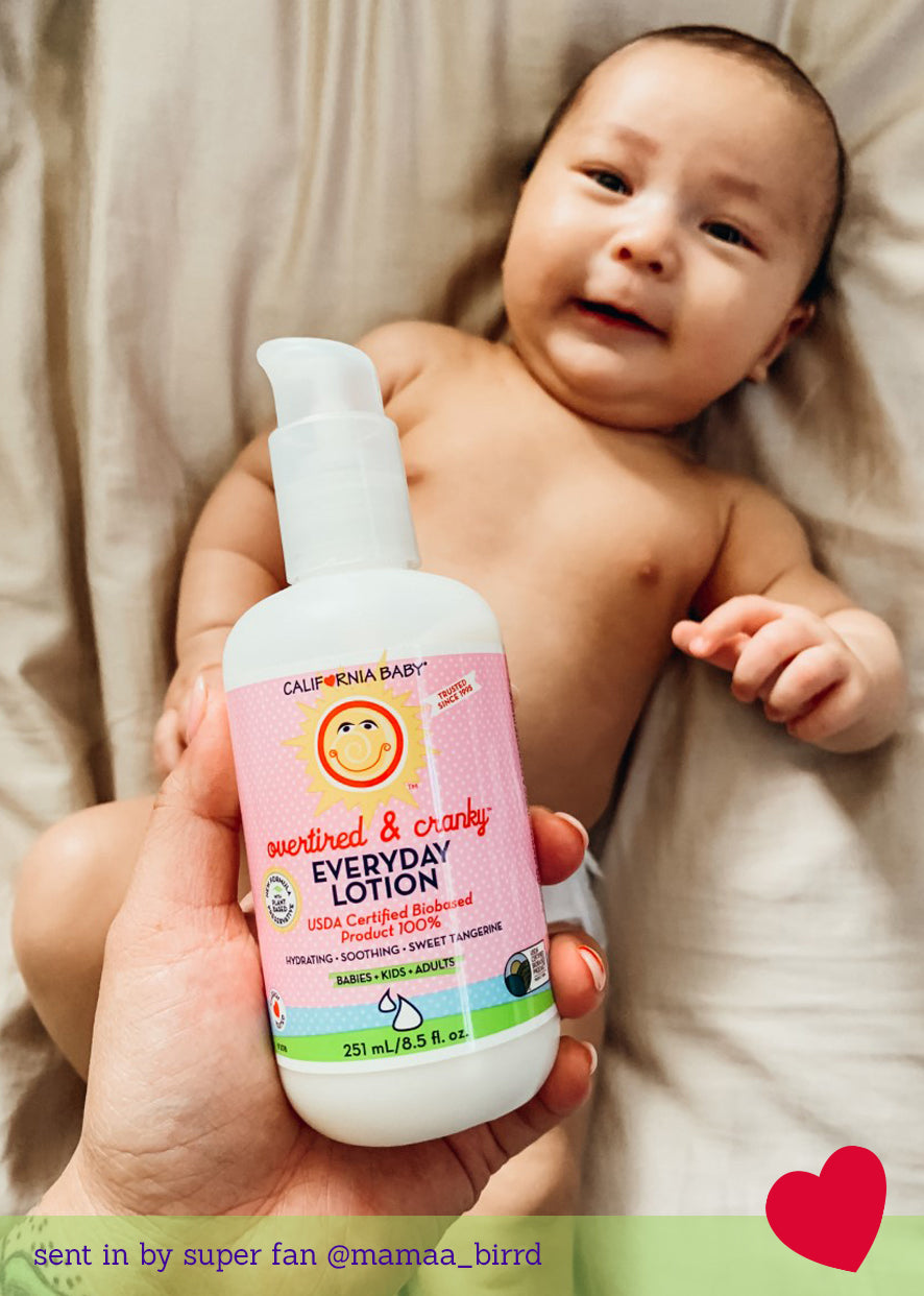 Hand holding lotion bottle with white pump and pink label; baby in background