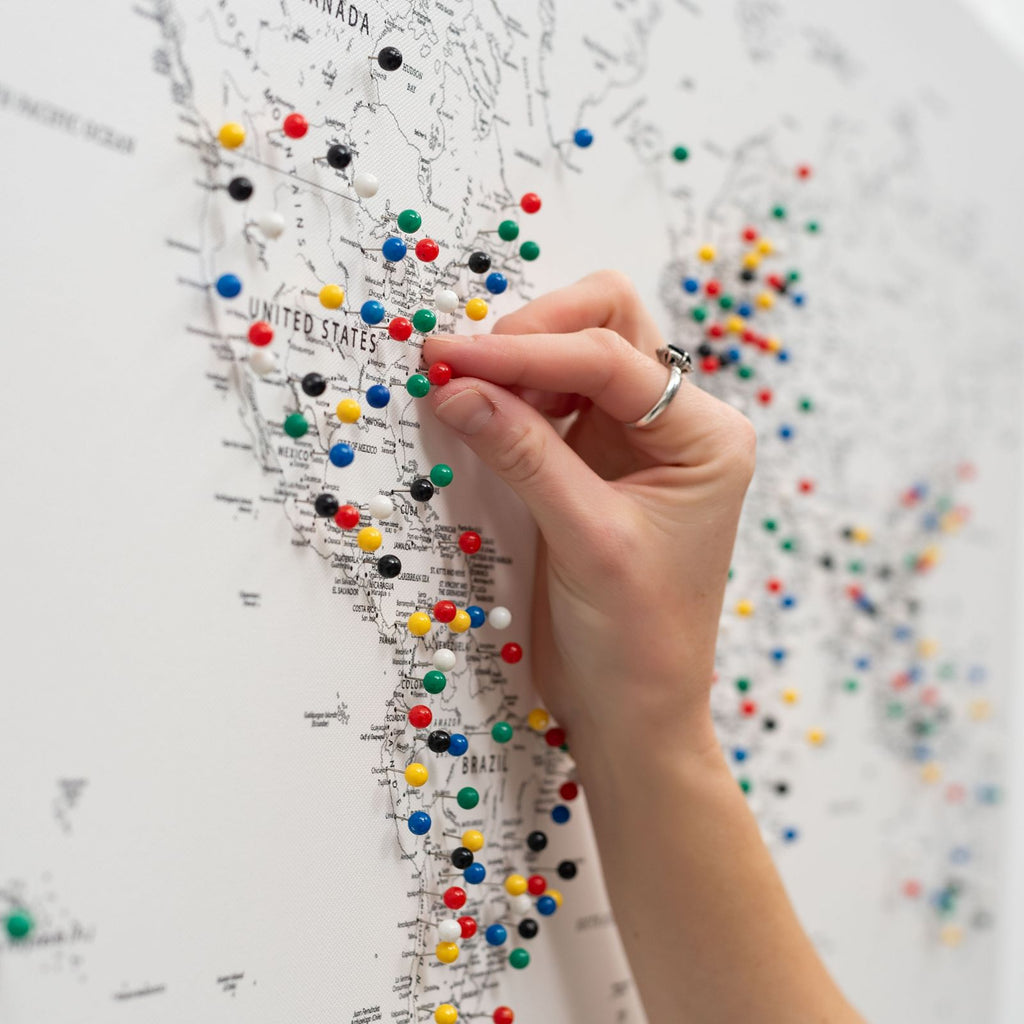Hand holding green, red, blue, and yellow push pins on a canvas world map