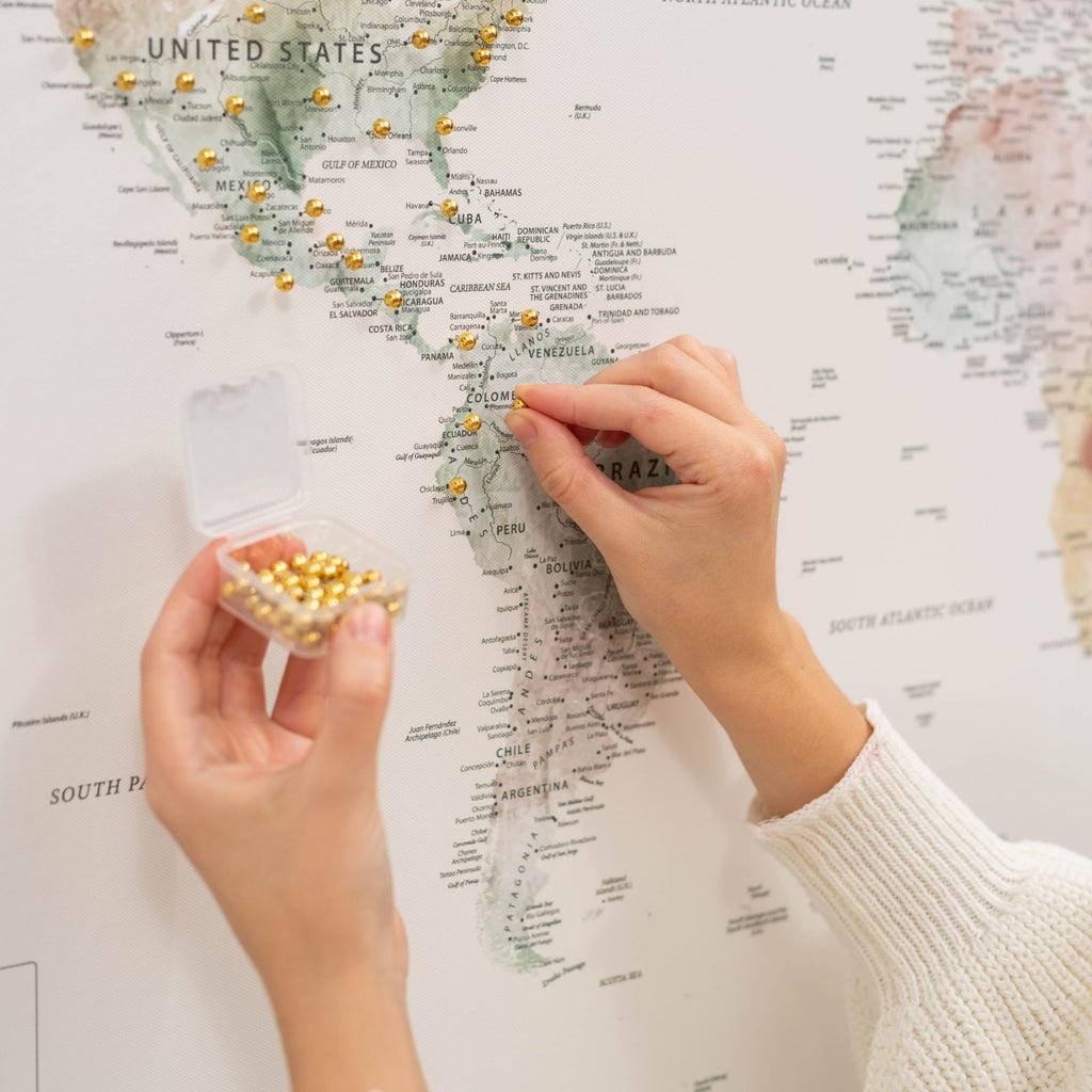 Hands placing gold push pins on a canvas world map; small pin container shown