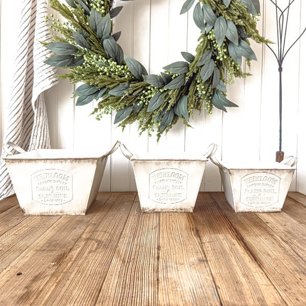 Three matching distressed white metal planters with handles lined on a wooden surface.
