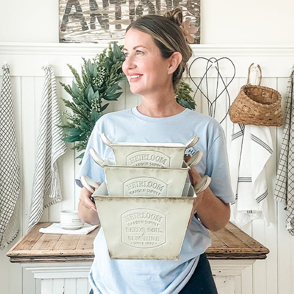 Woman holding three distressed white metal planters with handles and embossed garden seal.
