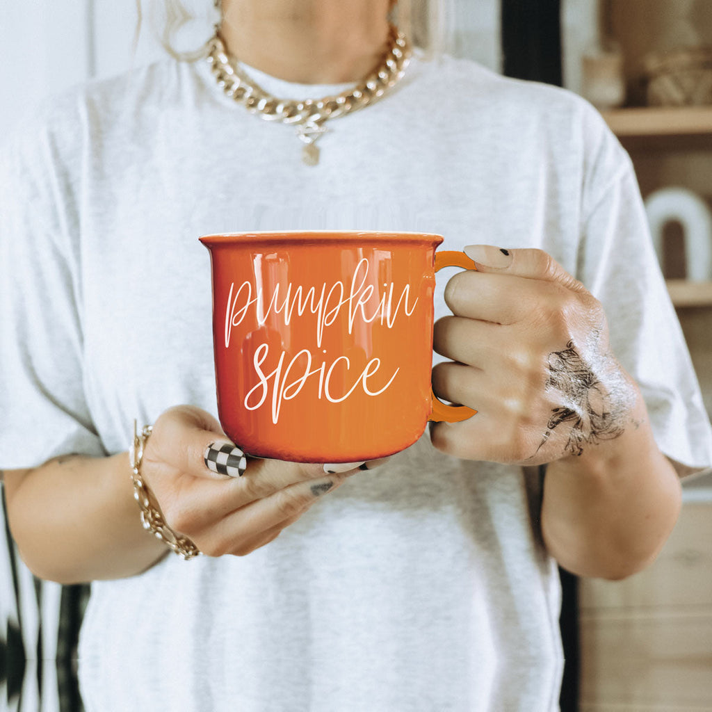 Orange ceramic mug with white cursive pumpkin spice text, held near a light shirt