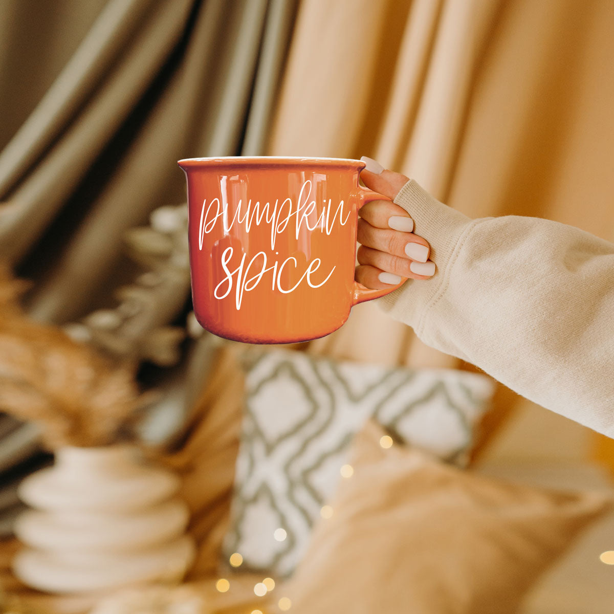 Orange ceramic mug with white cursive pumpkin spice text, held by a knitted sleeve