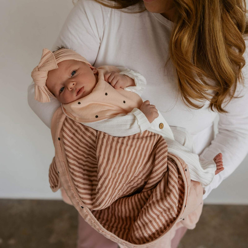 Blush muslin quilt wrap with striped layer held around a baby