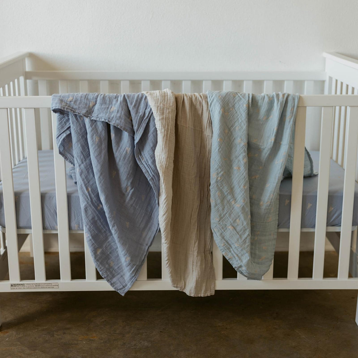 Crib display of three muslin swaddle blankets in light blue and beige, cloud prints