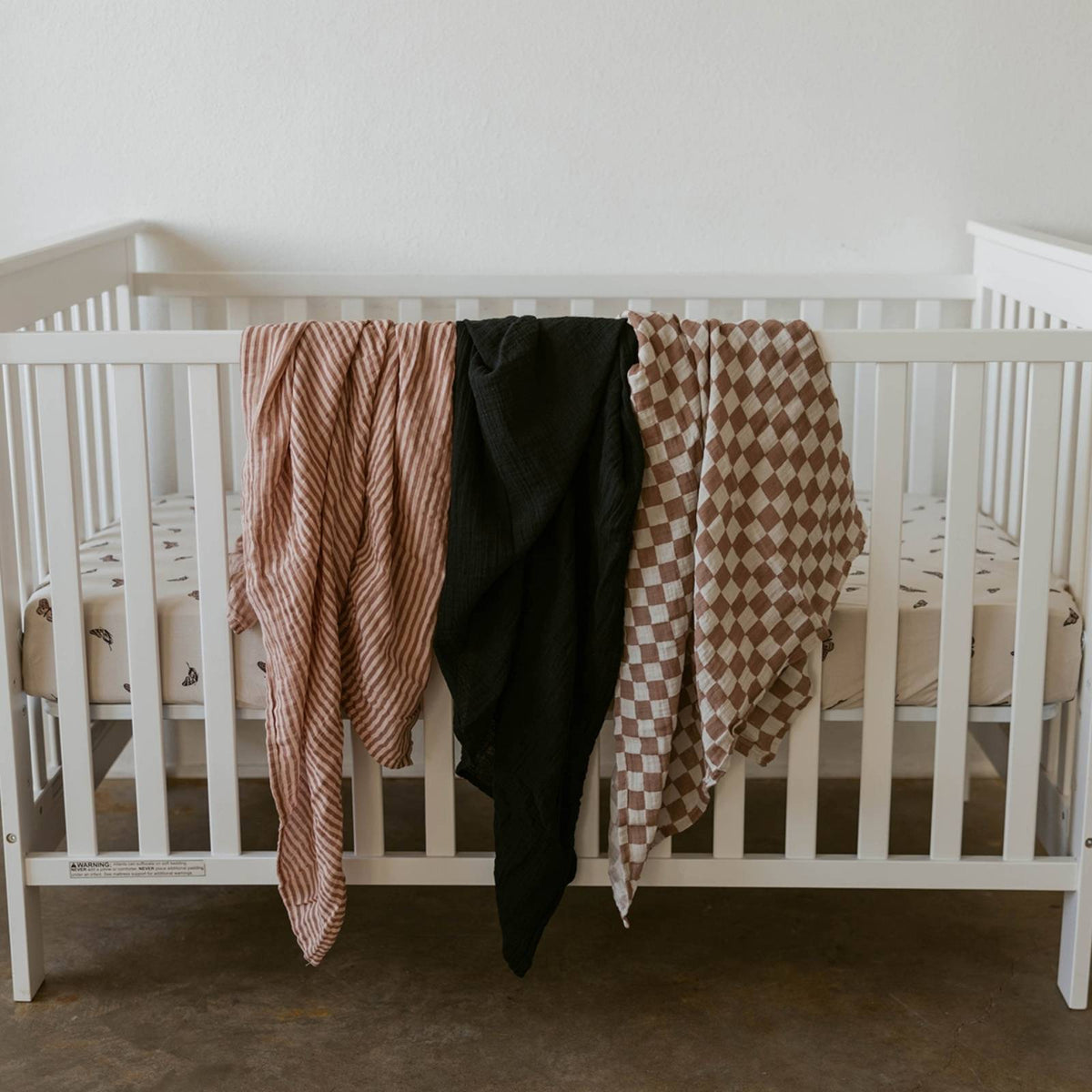 Three swaddles draped on a crib: rose stripe, dark solid, and brown checkered