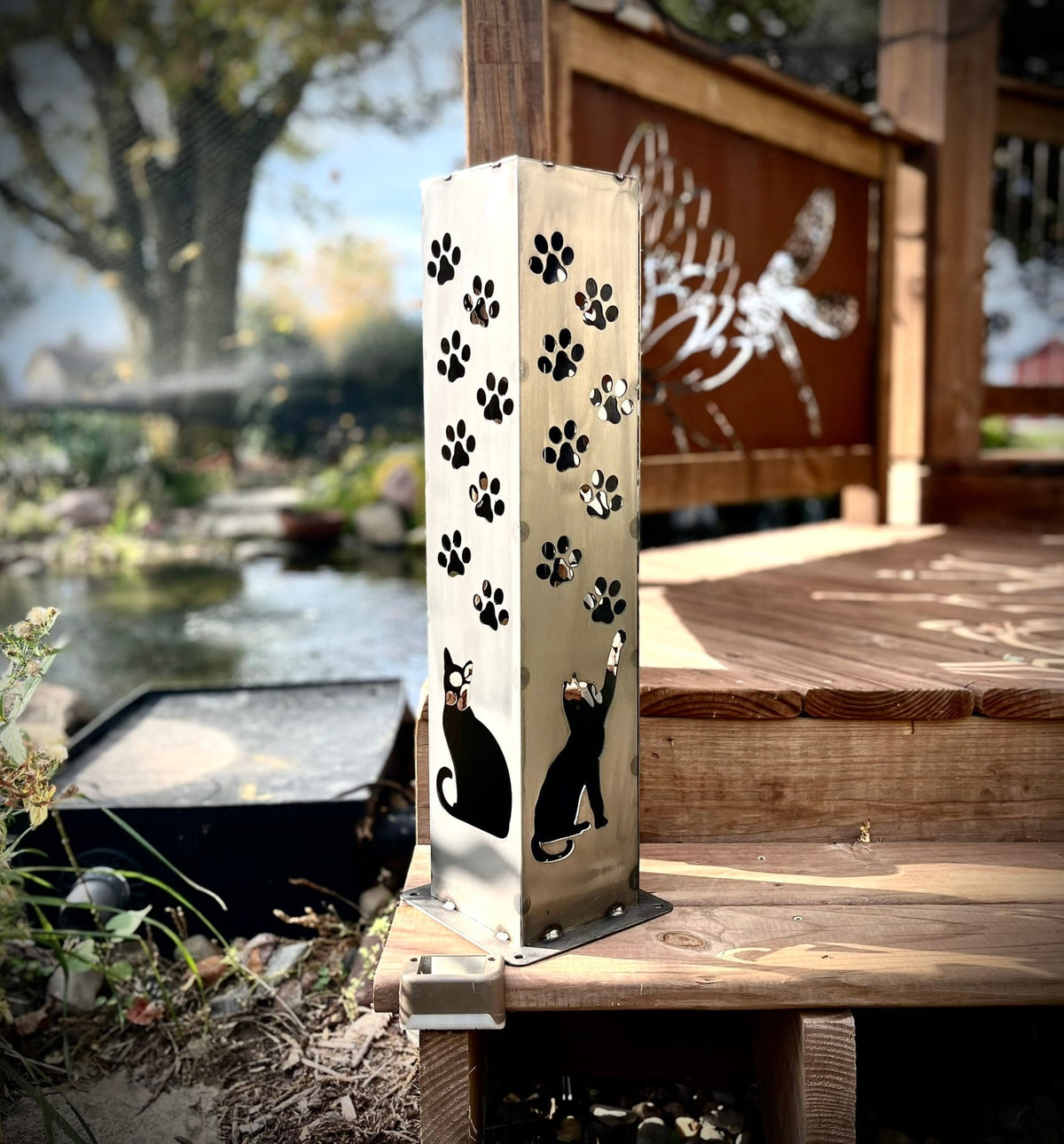 Brushed steel solar pillar with paw prints and dog-and-cat silhouettes beside pond