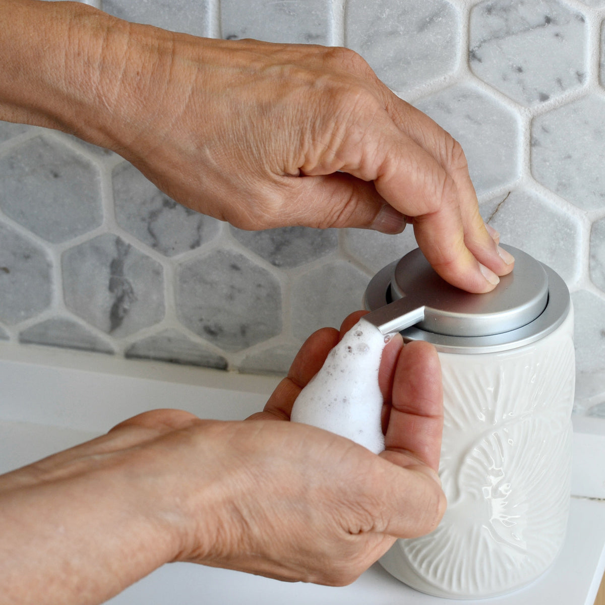 Hands pressing the top pump of a white ceramic foaming soap dispenser with spout