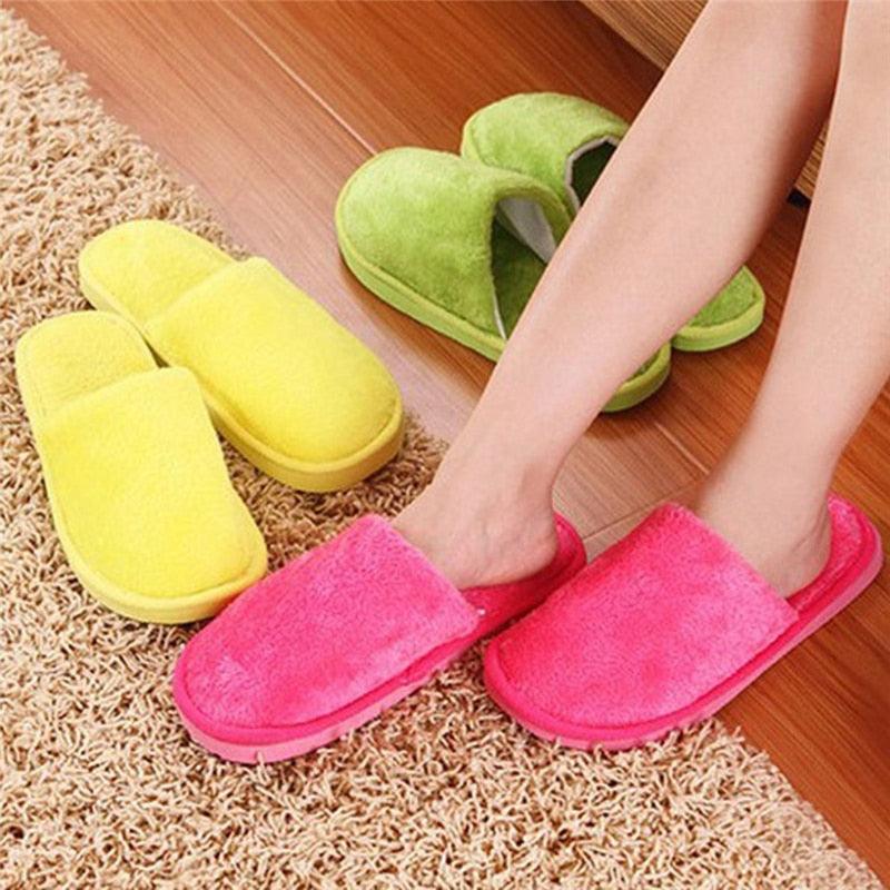 Pink slip-on fuzzy slippers on feet with yellow, green, and other pairs nearby