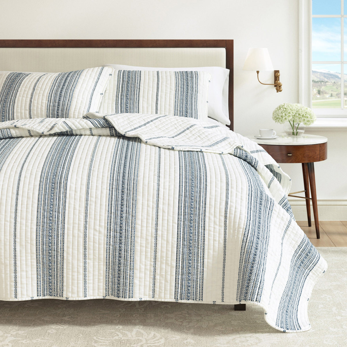 Striped quilt on bed with cream base and navy vertical stripe pattern.